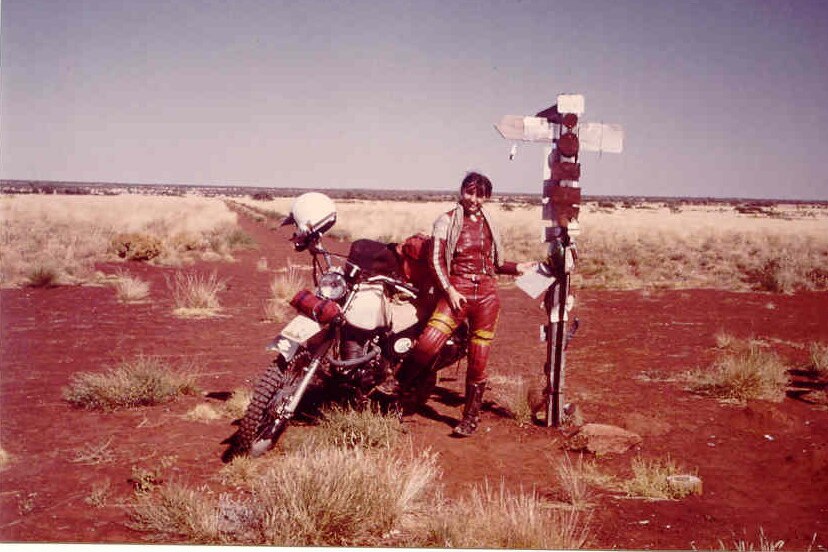 A woman standing in the middle of the desert with a motorbike.