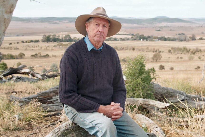 A farmer wearing a wide-brim hat and a wearing a sweater sits on his dry property