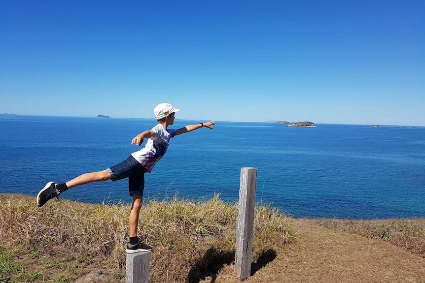 A boy stands on a pole in a ballet pose looking out to the ocean.