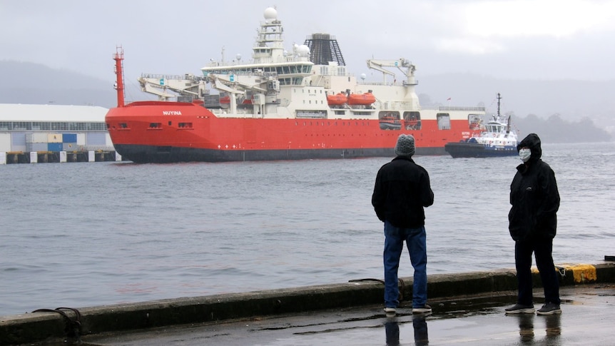 Two people, one wearing a mask, stand dockside next to a large ship