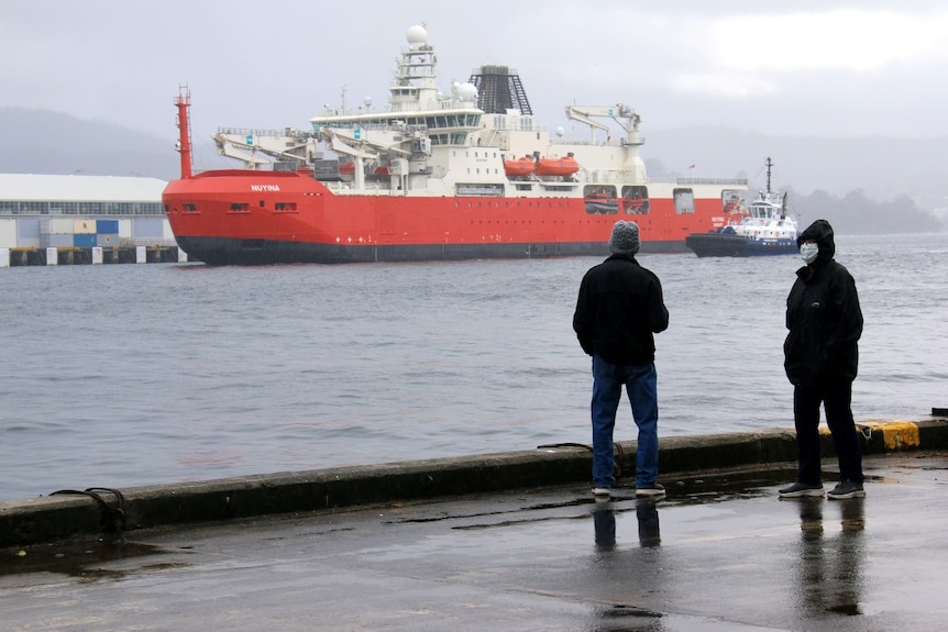 Two people, one wearing a mask, stand dockside next to a large ship