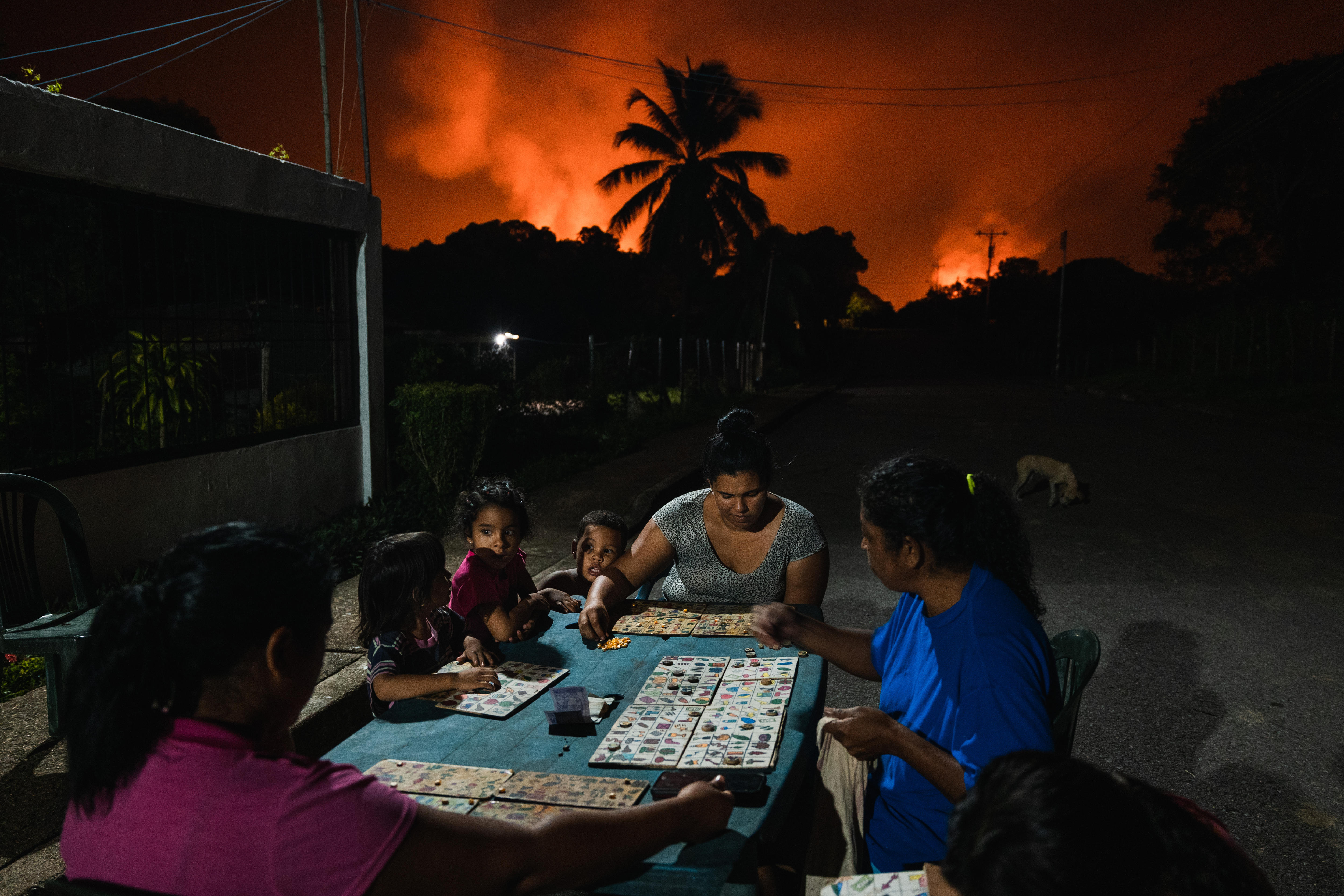 A family sits around an outdoor table playing a board game as a big bush fire rages behind them in the distance