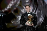Superstar footballer Lionel Messi stands smiling on a stage, looking down at the audience as he holds a trophy.