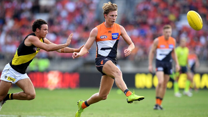 An AFL player kicks the ball downfield while being chased by an opposition player.