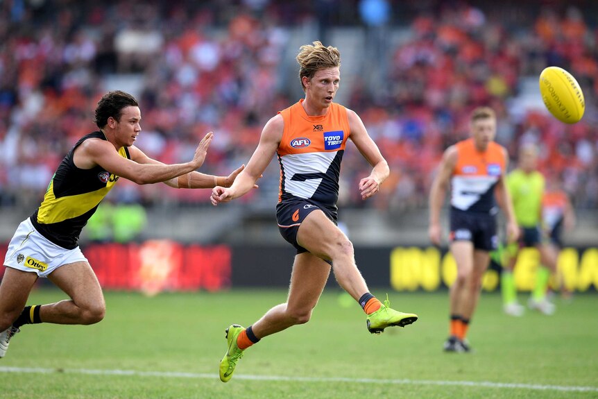 An AFL player kicks the ball downfield while being chased by an opposition player.