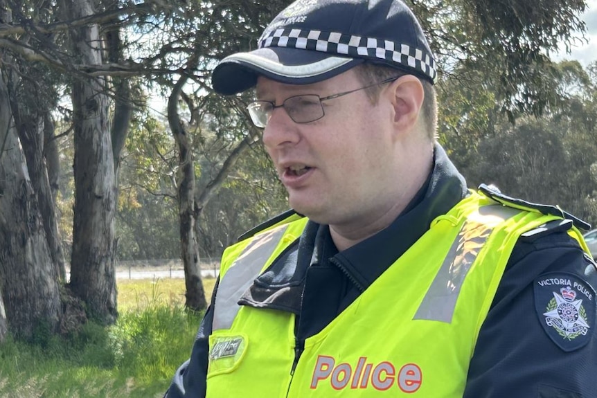 A policeman wearing high-vis stands near a road in the country and speaks to the media.