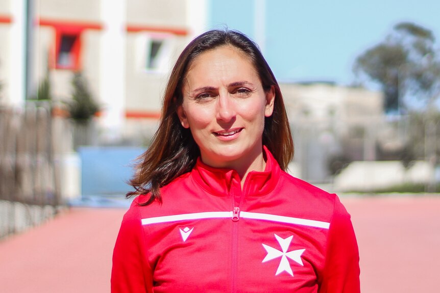 Mariella wears a red sports jacket with a white Maltese cross on the front, there's a blurred athletics track in the background