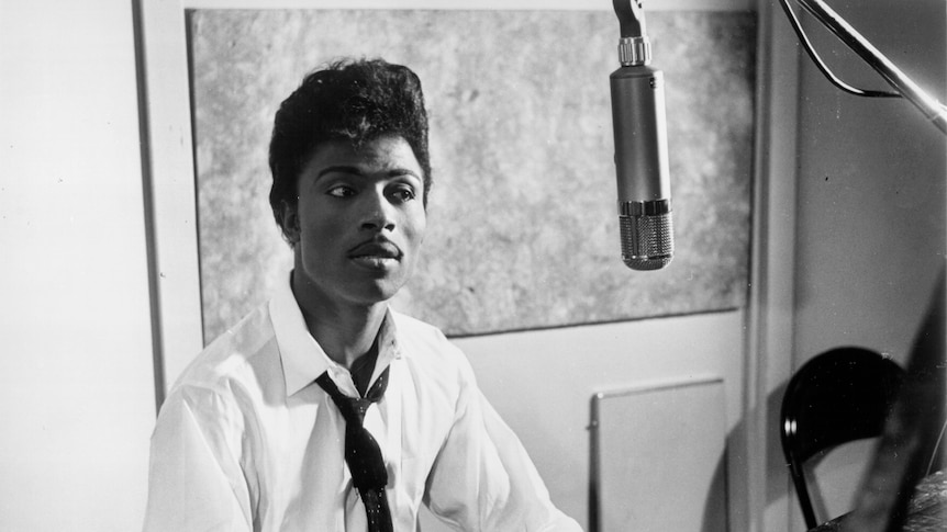 a black and white image of Little Richard in shirt and tie, playing the piano with a hanging microphone in front of him