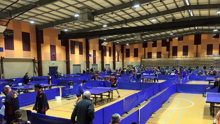 a wide shot of  a hall with many table tennis tables and players in action
