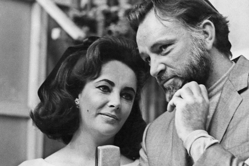 A black and white photo of Elizabeth Taylor and Richard Burton standing in front of a microphone