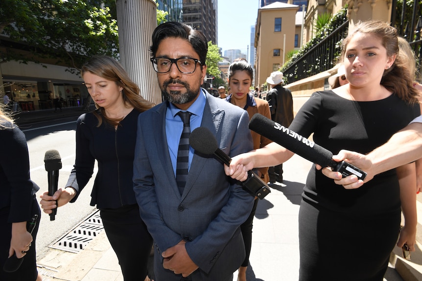 Man walking in suit surrounded by journalists holding microphones