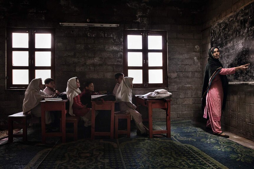 Children sitting at desks watching their teacher point to a lesson on the blackboard.