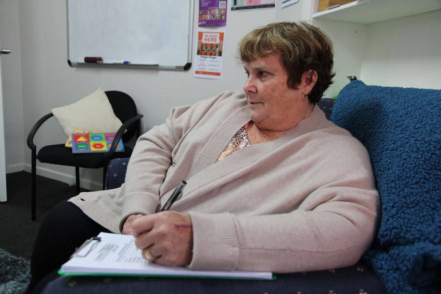A woman in a beige cardigan sits on a couch with pen and paper.
