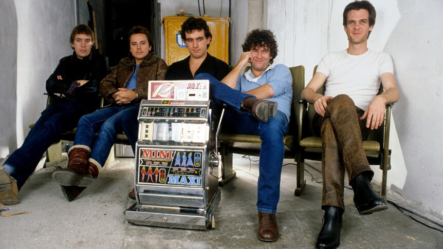  Five Cold Chisel band members sit in connected waiting room chairs. A pokie machine in front of them on the floor