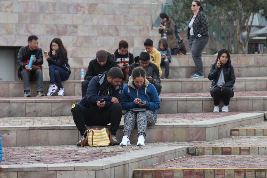 A number of people sit looking at their on the steps at Federation Square in Melbourne.