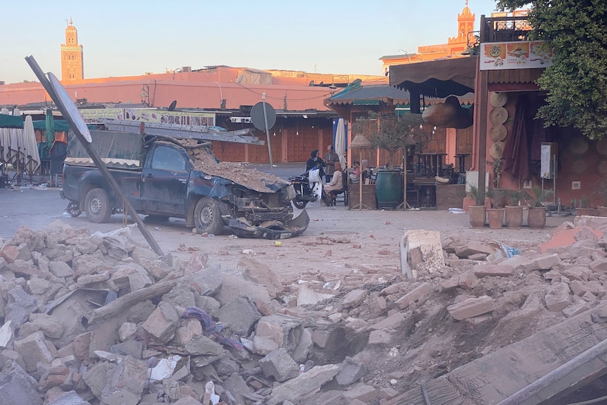 A dusty photo with rubble on the ground and some on a crushed pick up truck with people in the background