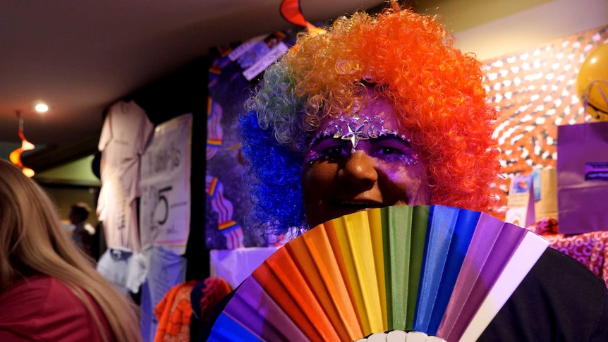 A person in a rainbow wig with a rainbow fan in front of their face.