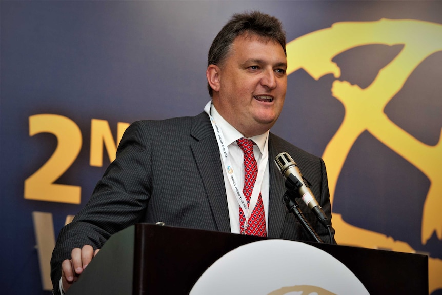 A man in a suit and tie speaking into a microphone at a lectern in front of a banner.