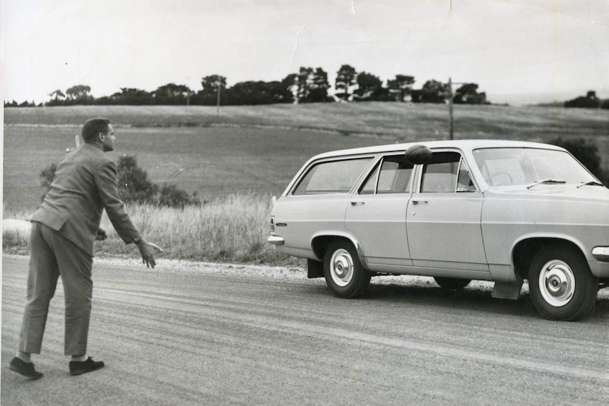 A black and white photo of a man handballing an AFL ball through a car window.