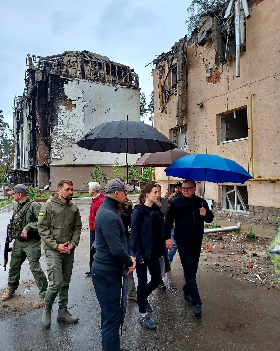 Sanna Marin walks with a group of people holding umbrellas, past partially destroyed buildings and rubble