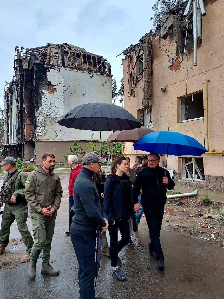Sanna Marin walks with a group of people holding umbrellas, past partially destroyed buildings and rubble