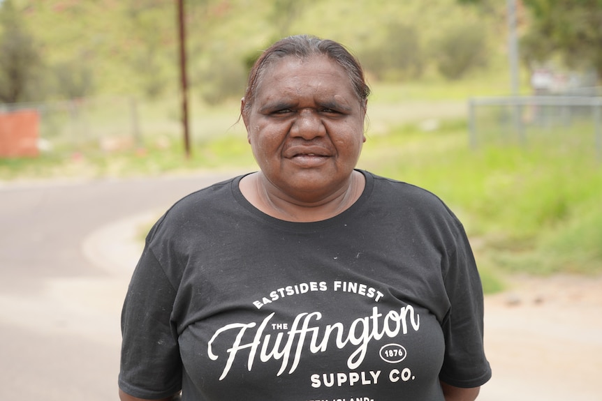 An Indigenous woman with her hair tied back, wearing a dark T-shirt, stands outside.