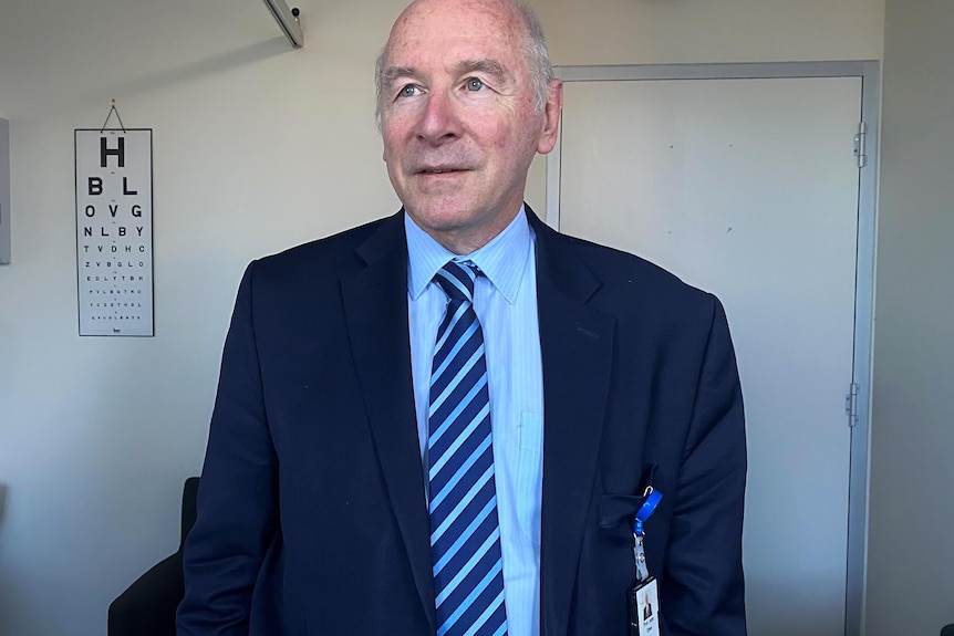 John Olver, who's wearing a blue shirt and navy jacket, poses for a photo in his office, an eye chart on the wall behind him