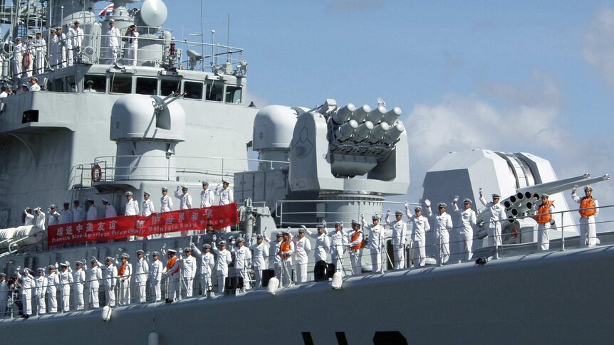 Looking from below, you look up at a large Chinese warship which has its deck rails lined by sailors in white uniform waving.
