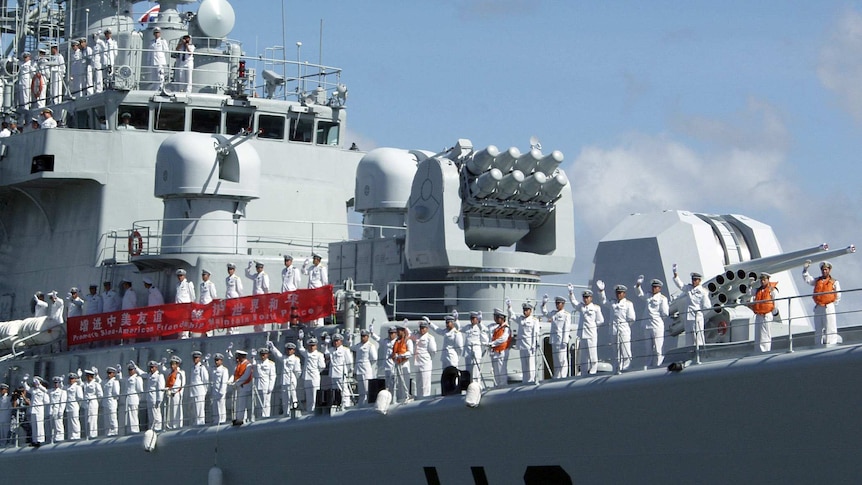 Looking from below, you look up at a large Chinese warship which has its deck rails lined by sailors in white uniform waving.