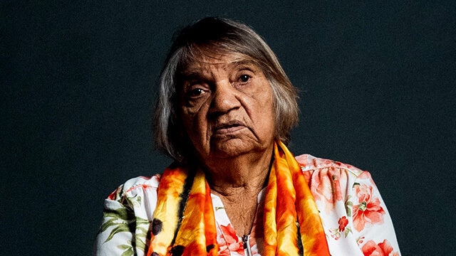 Aunty Muriel Bowie is a 78 year old Aboriginal woman sitting with a colorful scarf and a neutral expression.
