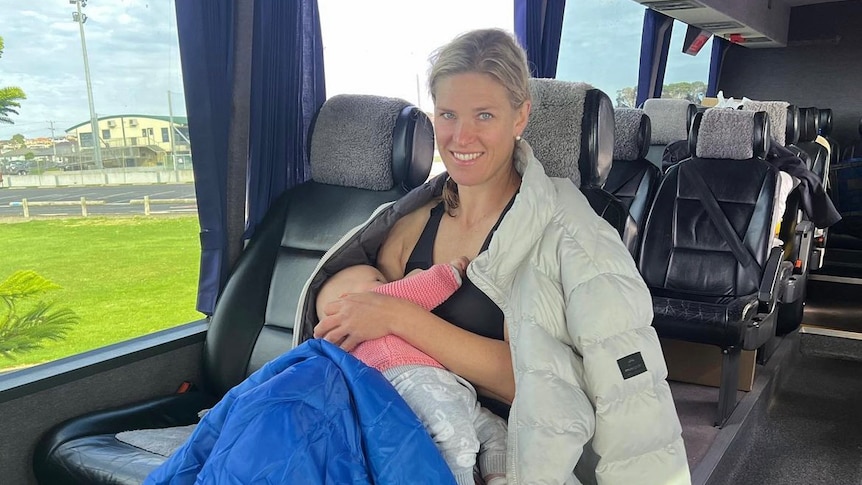 Kate Næss is sitting on a bus, she has a jacket draped around her shoulders and is breastfeeding a baby.