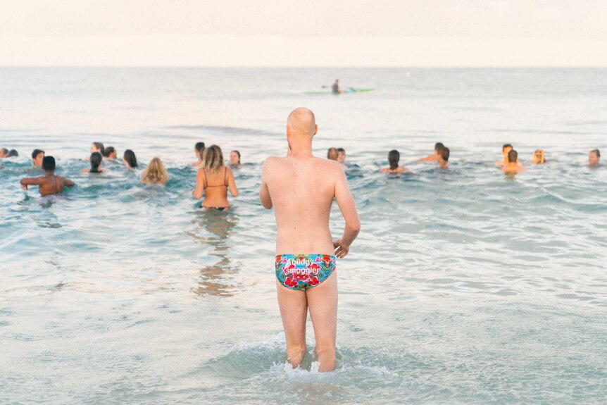 A man wearing a budgie smuggler swimwear looks out towards people swimming in the ocean