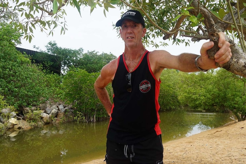 A man in a black hat and black singlet stands against a tree at the Little Mindil estuary.