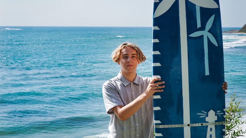 A boy holding a painted surfboard with the ocean behind him.