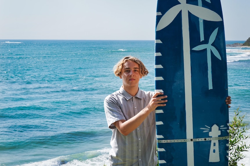 A boy holding a painted surfboard with the ocean behind him.