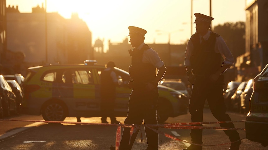 Two police officers stand by police tape on a road as the sun rises in the background casting an orange hue over the photo