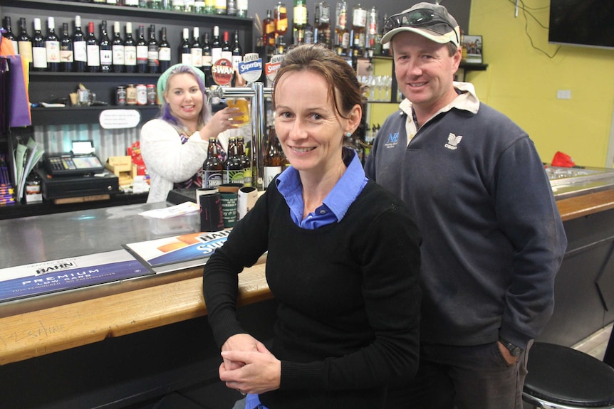 A woman pours a beer behind a bar in a country pub and a woman and a man stand in the foreground of the photo.