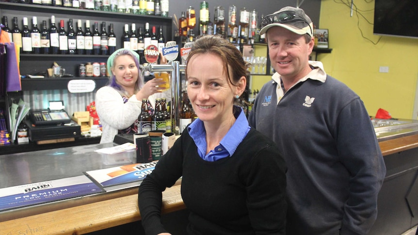 A woman pours a beer behind a bar in a country pub and a woman and a man stand in the foreground of the photo.