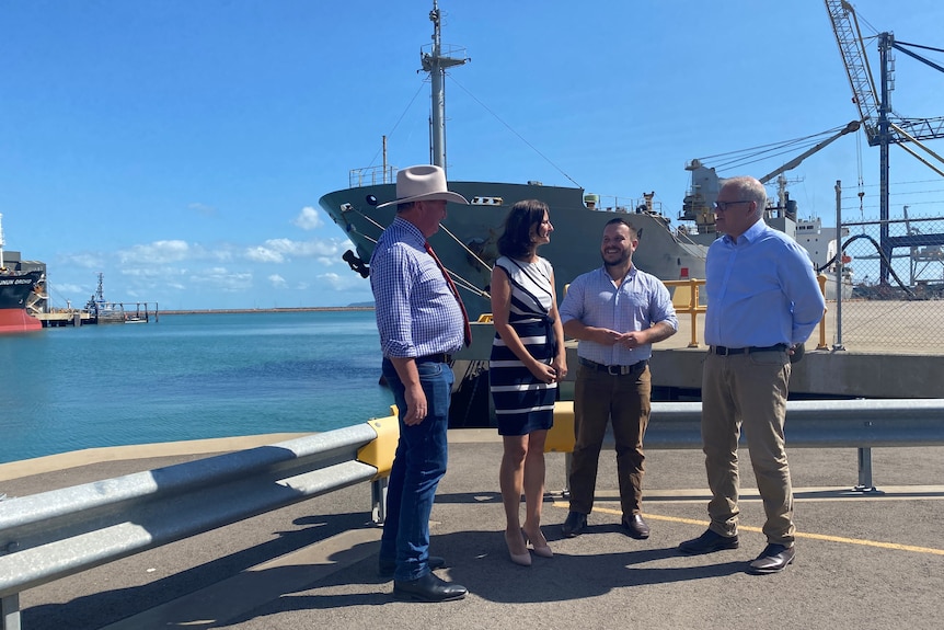 Politicians, three men, and TEL chief executive, a woman, gather in front of a ship with water behind. 