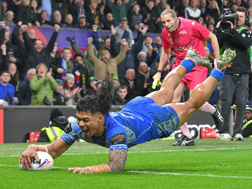 A Samoan rugby league player dives over the line with a smile on his face as a referee and a cameraman watch in the background.