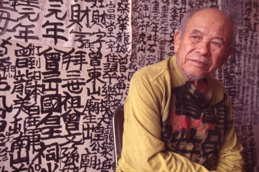 An older Hong Kong man sits looking into distance, behind him is a black ink calligraphy decorated wall.