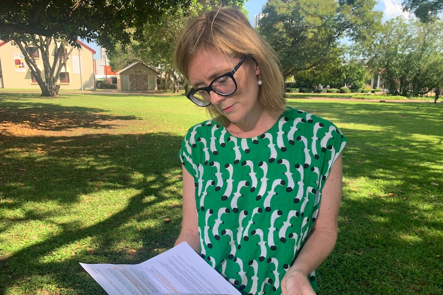 A woman in a green patterned dress reading through documents and standing in a park.
