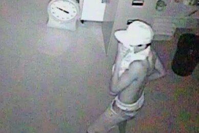 A black-and-white CCTV image of a young man indoors in a white hat and covering his face.