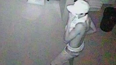 A black-and-white CCTV image of a young man indoors in a white hat and covering his face.