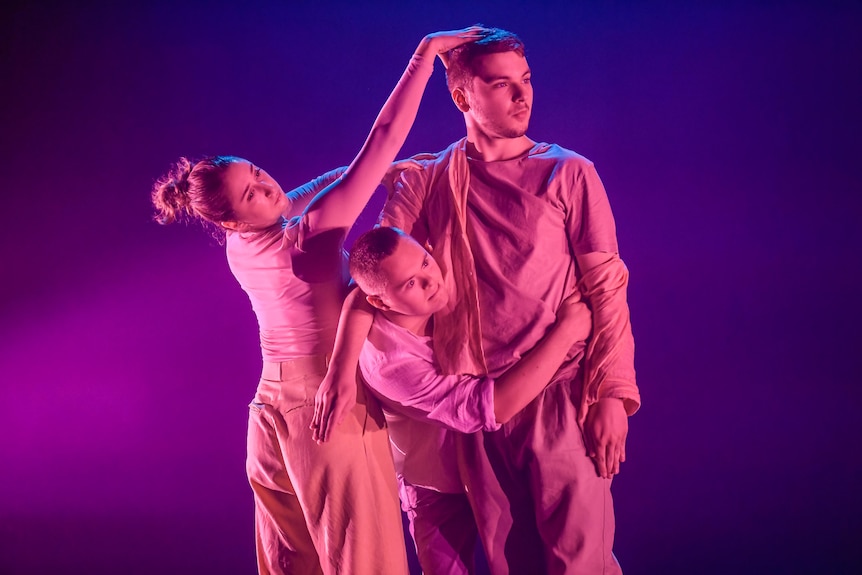 A white woman, a white man with down syndrome and another white man dance in loose fitting clothing on a stage.