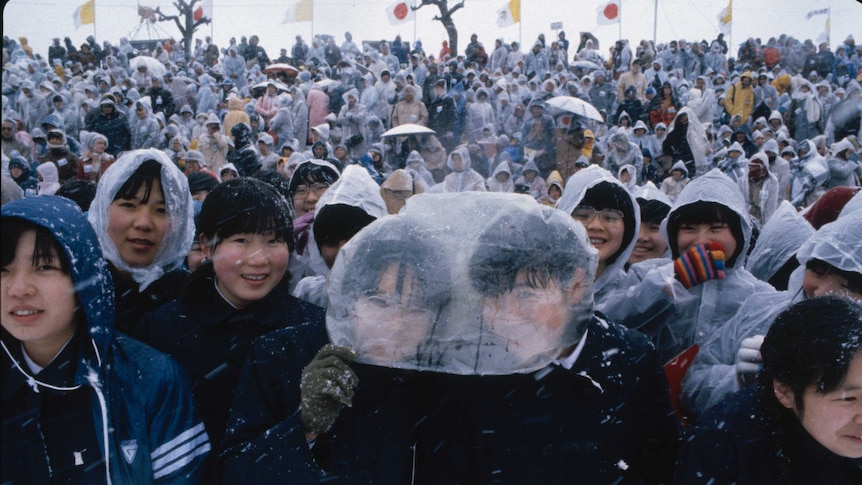 Two young Japanese students share a plastic bag as a head covering in a crowd in the snow.