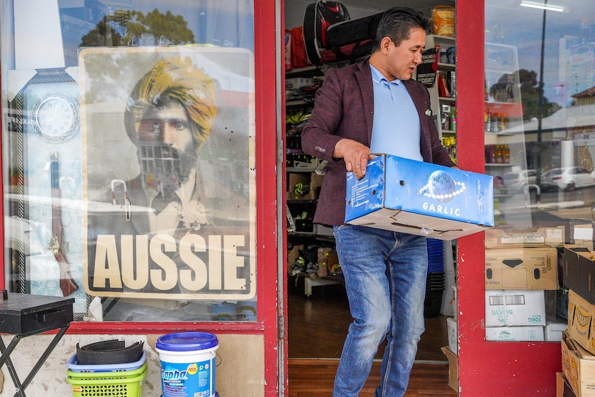 A man wearing jeans and a casual coat jacket walks out of a shop door carrying a cardboard box.