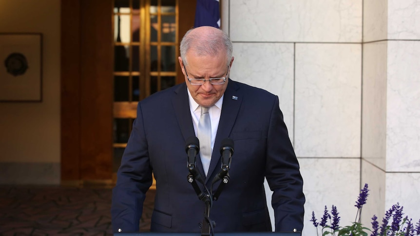 Scott Morrison looks down while standing at podium with Australian emblems on them in a courtyard with Australian flags