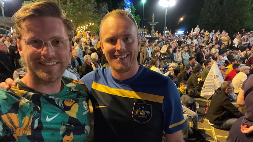 Two men in different Socceroos guernseys with a crowd behind them at night.