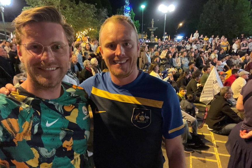 Two men in different Socceroos guernseys with a crowd behind them at night.